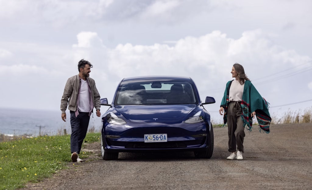 A man on the left and a woman on the right of a blue Drive Car Hire Tesla. They've stopped on a gravel road with grass and dandelions, the water in the background, and a bright, cloudy sky.
